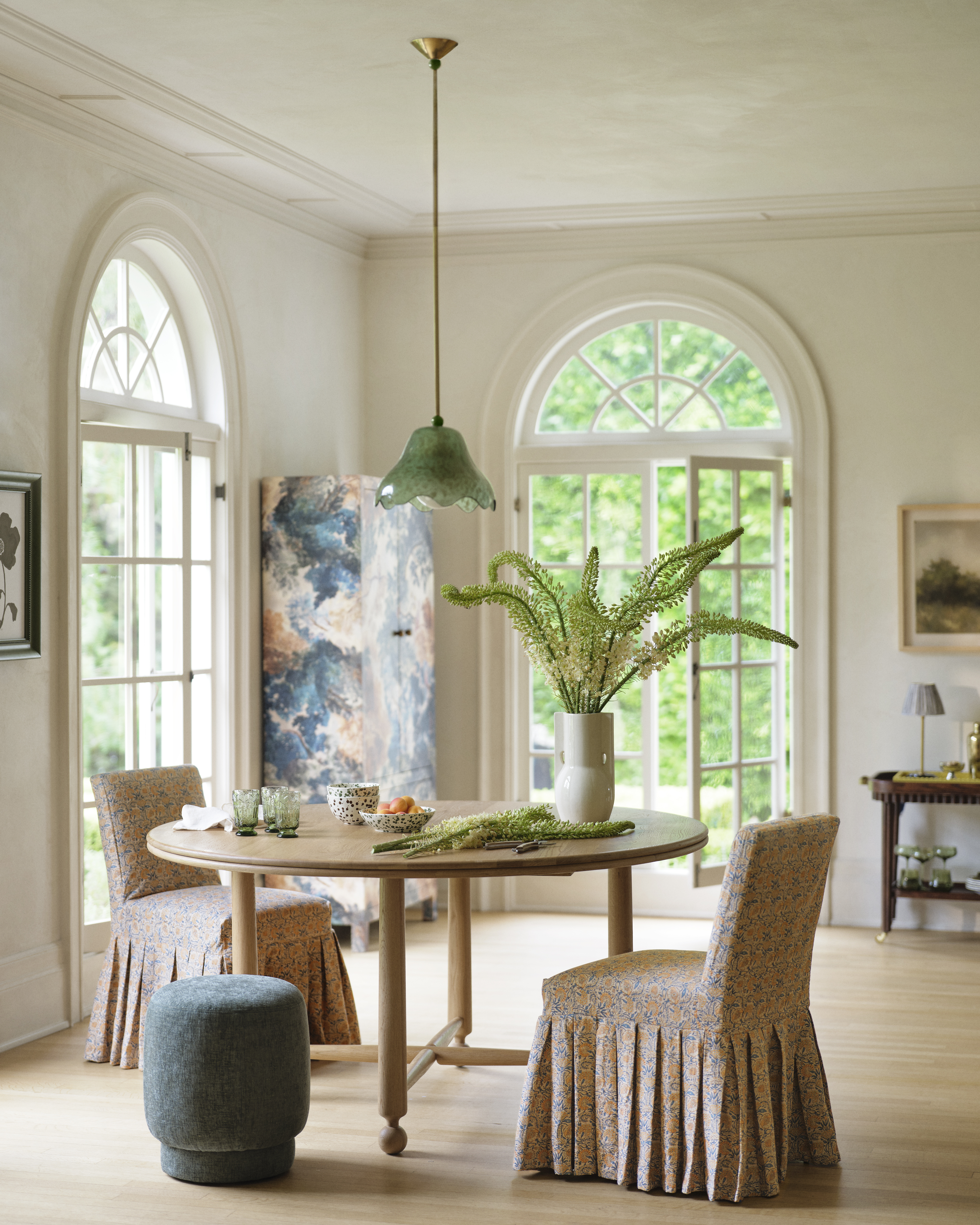 Green scalloped pendant light hanging in a sunlit dine-in kitchen that features a wooden table, skirted dining chairs, and a tapestry patterned room divider