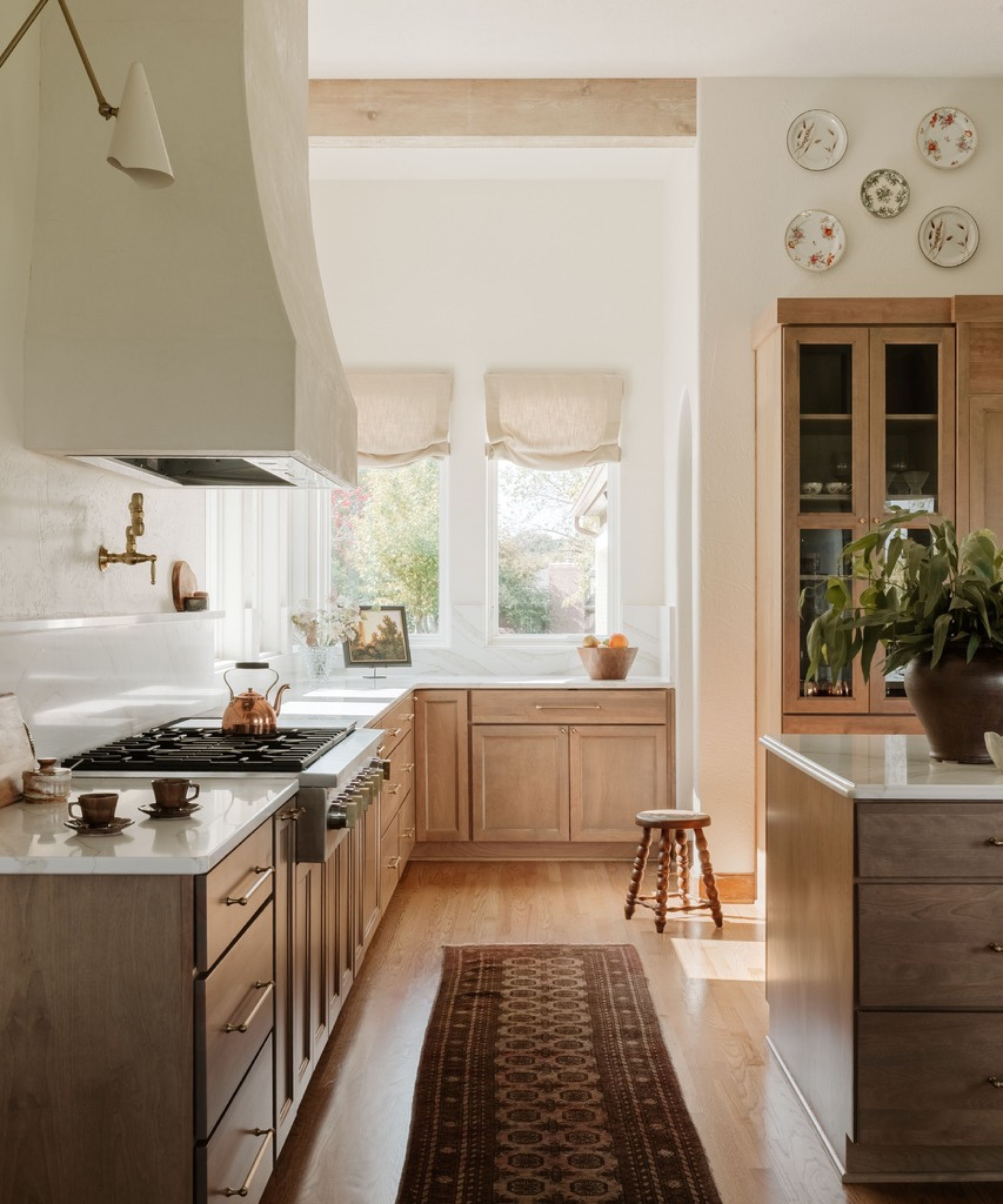 A sunlit kitchen with light wood cabinets, a plaster-finish range hood, and a long vintage runner rug on the hardwood floor leading toward large windows