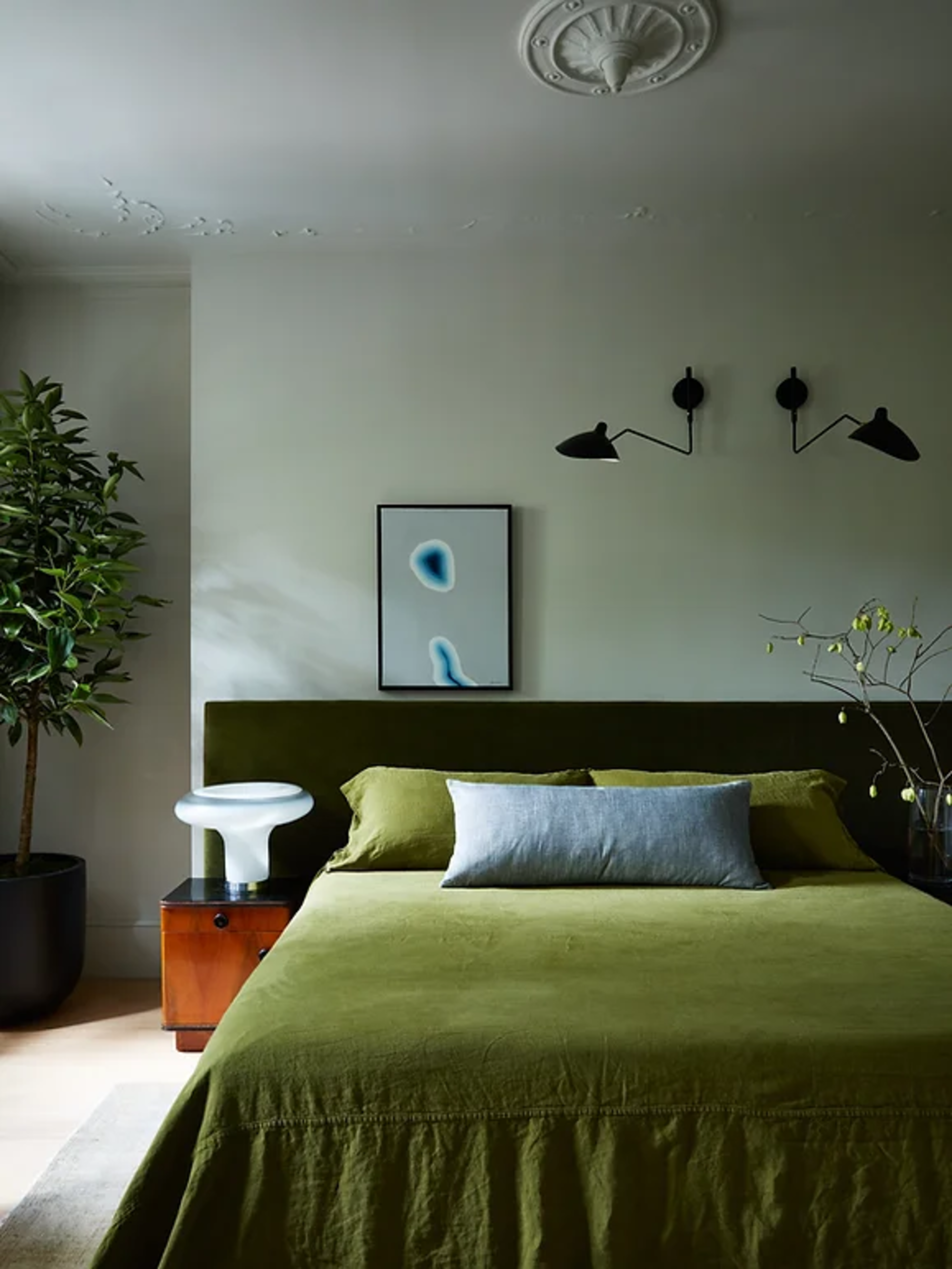 modern bedroom with sage green walls, deep green bedcover, velvet green headboard, artwork, plant in corner, and timber side table with light
