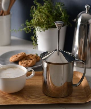 A modern stainless steel milk frother with cup of coffee in white porcelain cup and side of chocolate chip cookies on wooden-effect serving tray