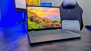 A sleek laptop on a wooden desk displays a vibrant landscape of mountains and flowers under a sunset. Nearby, a gaming chair and monitor create a cozy setup.