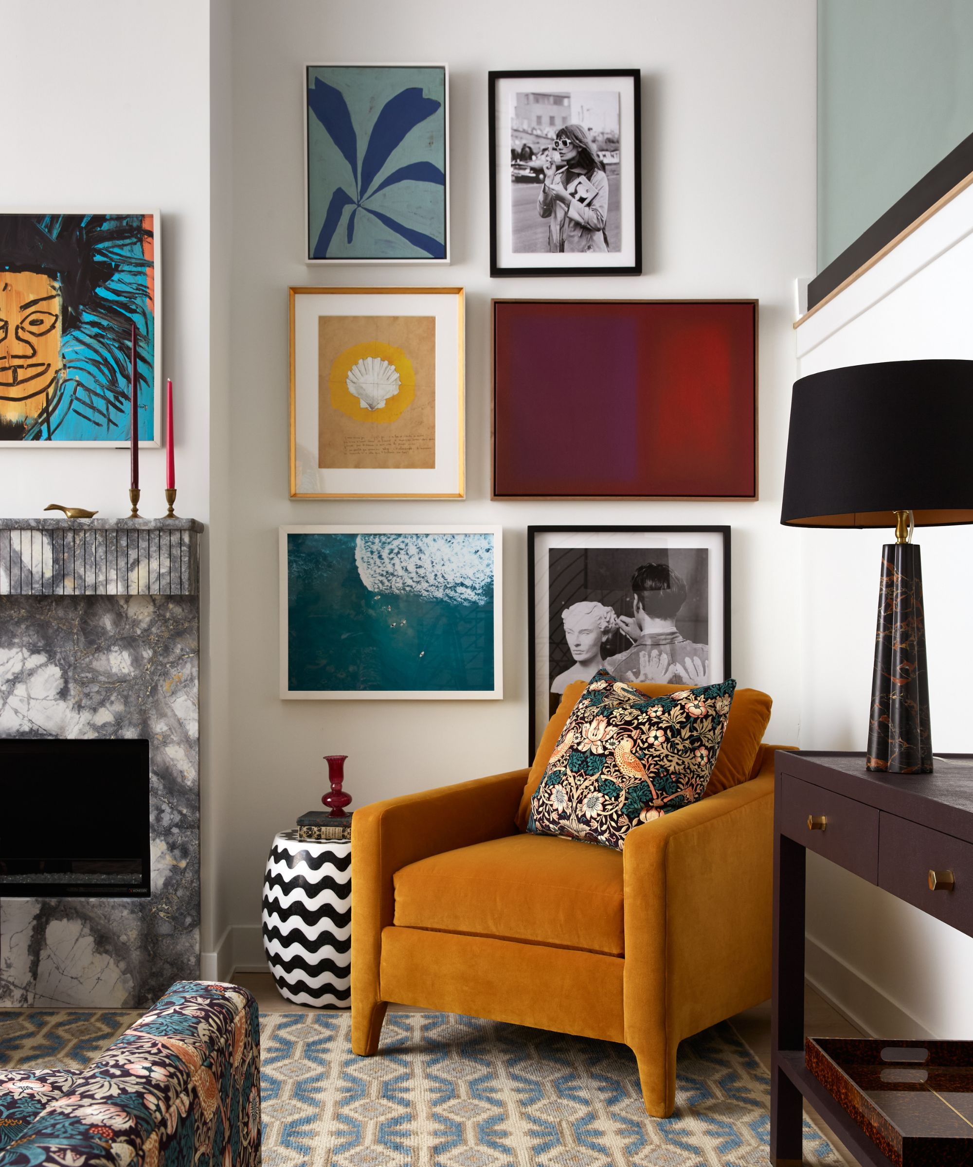 A modern living room with white walls, a gallery wall with colorful artwork, an ochre accent chair with a teal floral cushion, a pale blue and cream patterned rug, and a marble fireplace.