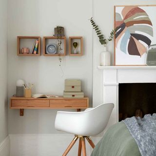 Bedroom with wooden desk and square shelves on the wall next to white chair and fireplace.