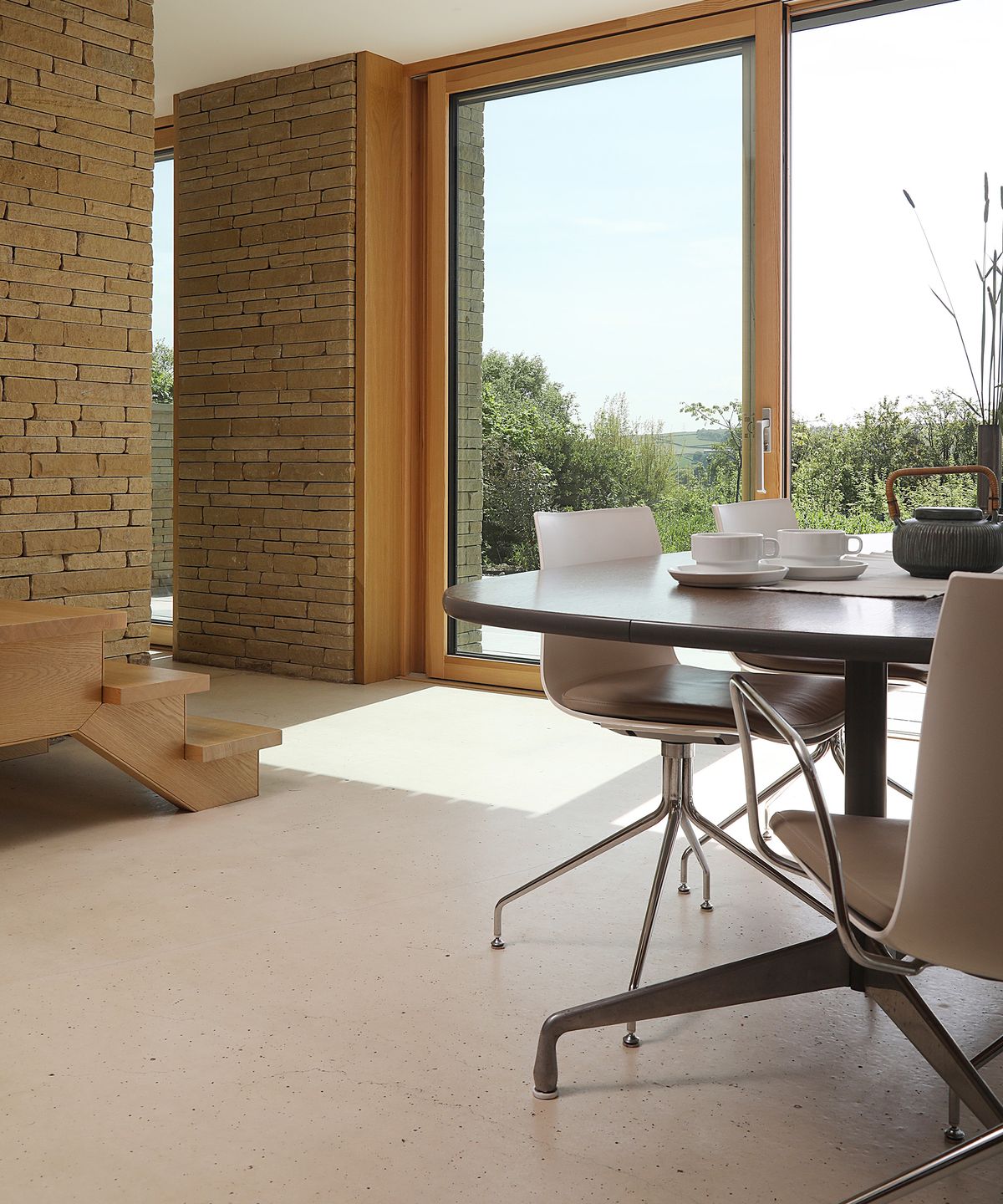 A contemporary living room with light coloured flooring a a sleek dining table. The walls are exposed brick with large glazed sliding doors leading outside