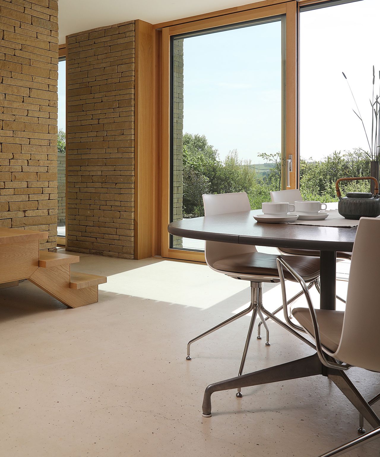 A contemporary living room with light coloured flooring a a sleek dining table. The walls are exposed brick with large glazed sliding doors leading outside