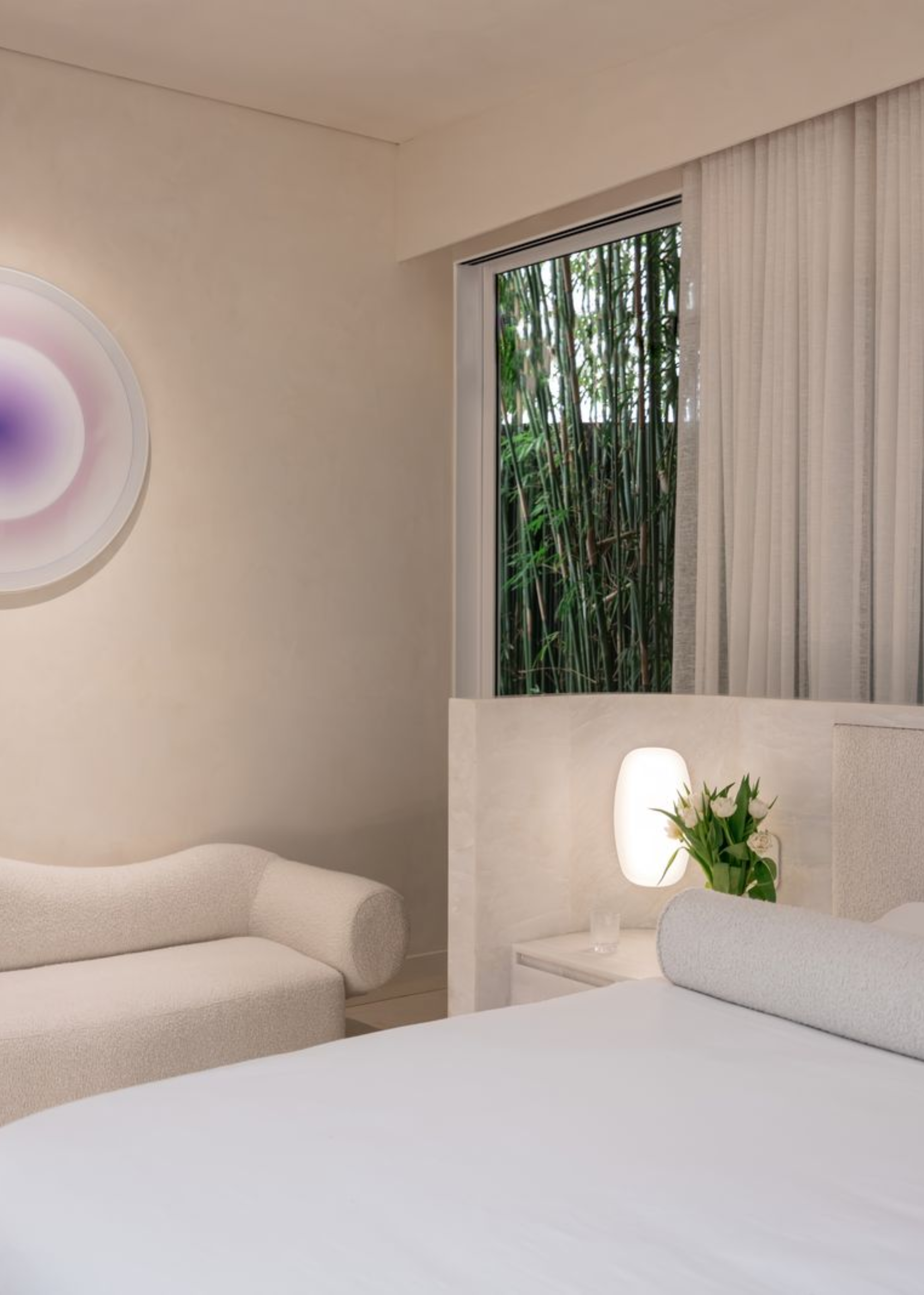 An off white bedroom with a cozy sofa, a purple wall sconce and a short headboard