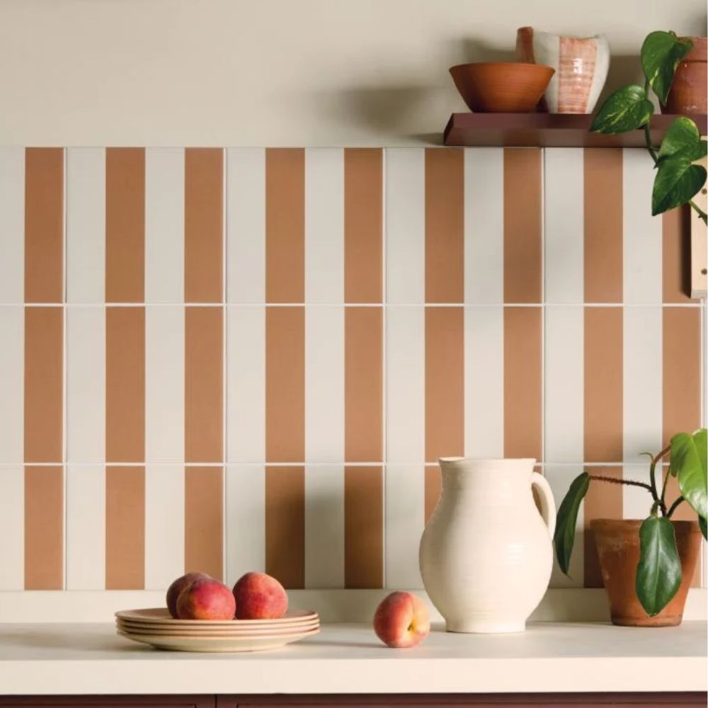 A kitchen splashback featuring alternating vertical terracotta and cream rectangular tiles creating a bold stripe effect. A cream ceramic jug, stacked plates with fresh peaches, and a potted plant are styled on the white worktop below, with a dark shelf holding bowls above.