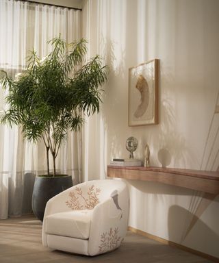 A white room with an embroidered armchair, a table built into the wall, and a potted tree in the corner