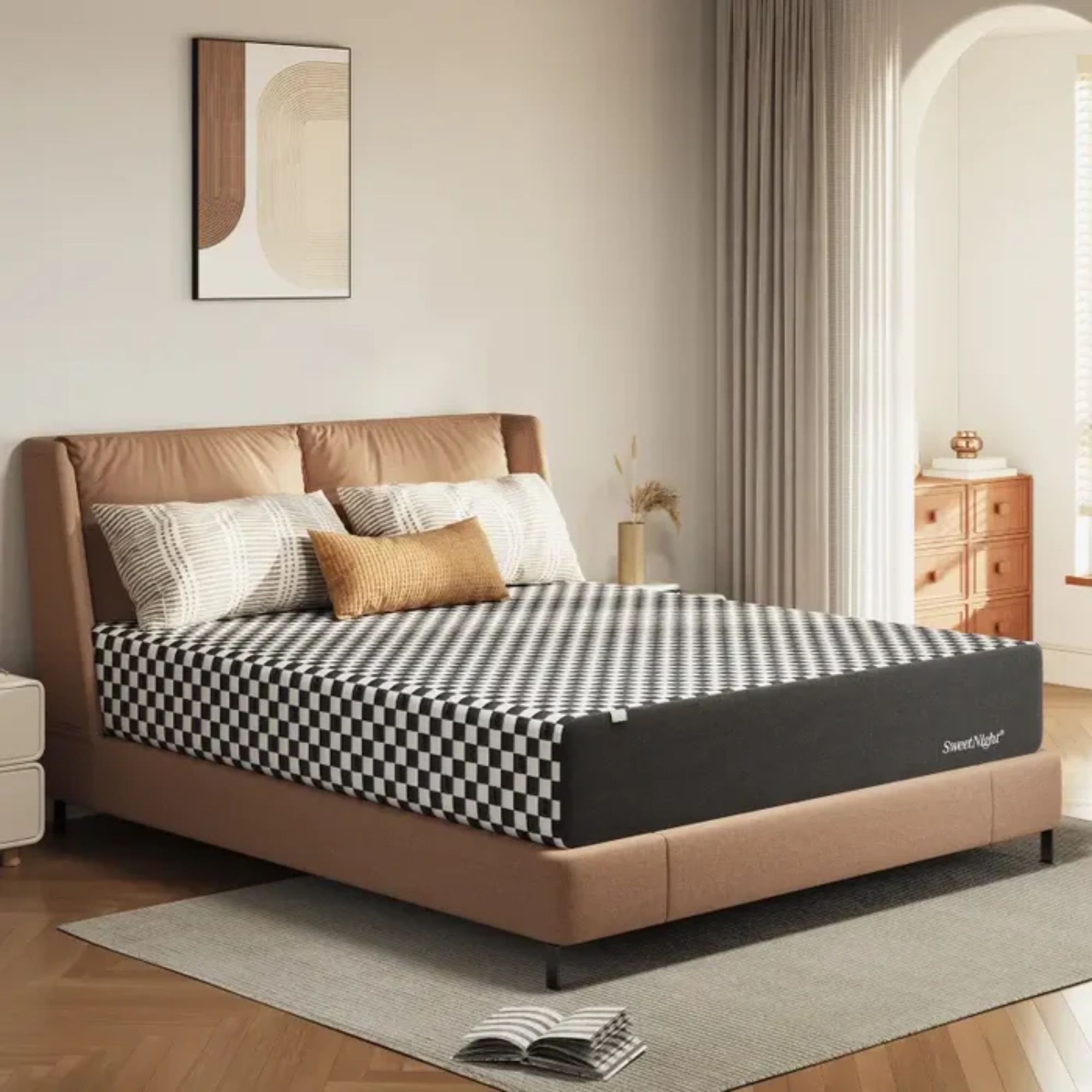 A black and white chequered SweetNight Prime Memory Foam Mattress pictured on a light brown upholstered bed in a cozy bedroom. On top of the bed are two patterned black and white pillows, and a rectangular burnt yellow cushion. Beneath the bed is a large grey area rug, with an open book pictured on it, and the floor is wood. To either side of the bed are modern white bedside tables, and the one on the right also features a gold vase with dried plants in it. Above the bed is a minimalist neutral artwork, and the walls are cream. To the right of the bed is an open curtain, with a smaller room section seen behind with a wooden chest of drawers and small gold vase.