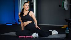 Woman in gym in 90/90 position on floor. She wears black sportswear