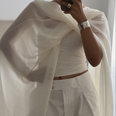 A woman taking a mirror selfie in a crisp white top, shorts and sheer white cape