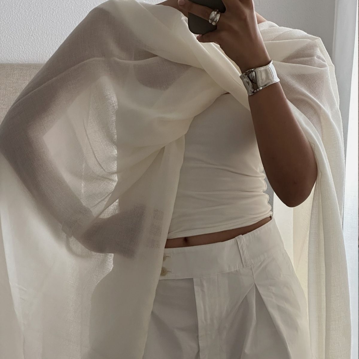 A woman taking a mirror selfie in a crisp white top, shorts and sheer white cape
