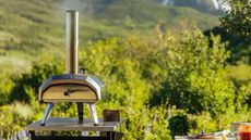 The Ooni Karu 12 pizza oven on a stainless steel table, outside, in front of a mountain 