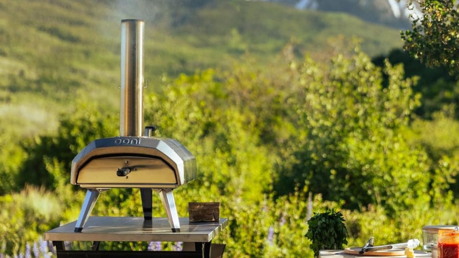 The Ooni Karu 12 pizza oven on a stainless steel table, outside, in front of a mountain 