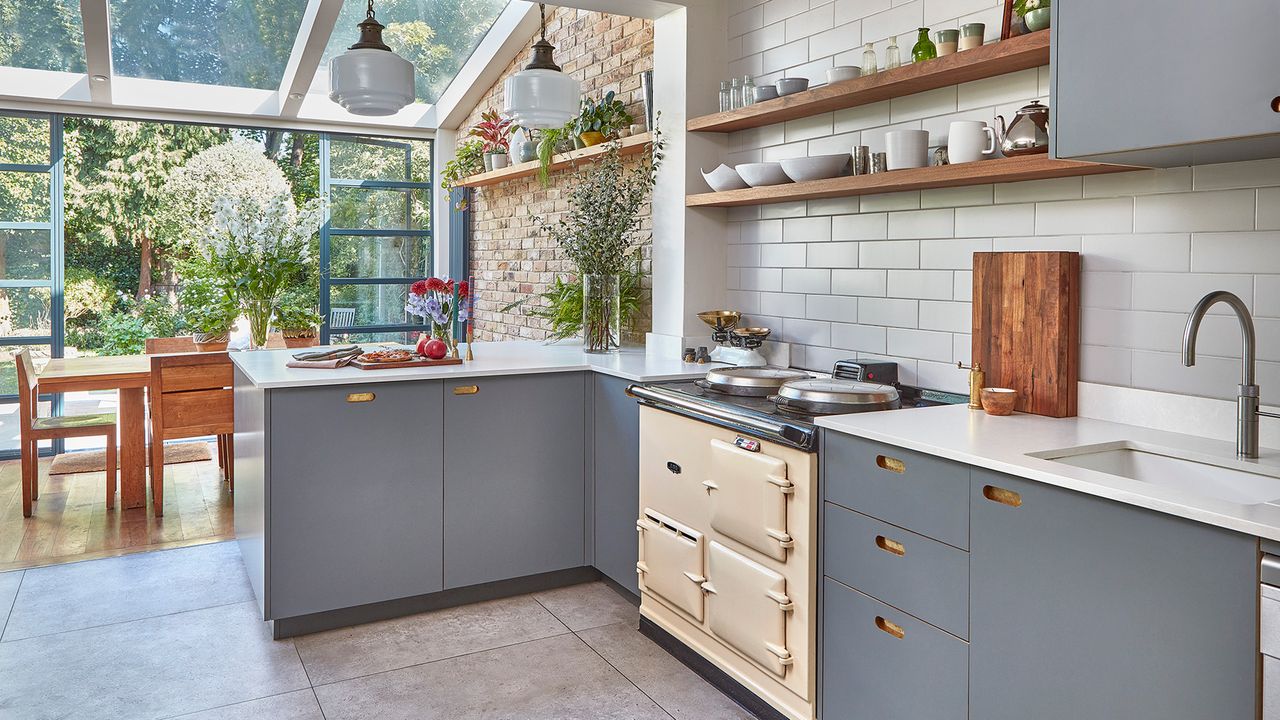 A modern traditional kitchen diner with blue cabinetry, cream aga cooker, wooden shelving, white pendant fixtures and wooden dining set