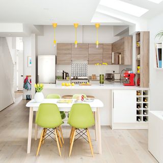 A white kitchen with wooden cabinets, yellow pendant lights and green dining chairs