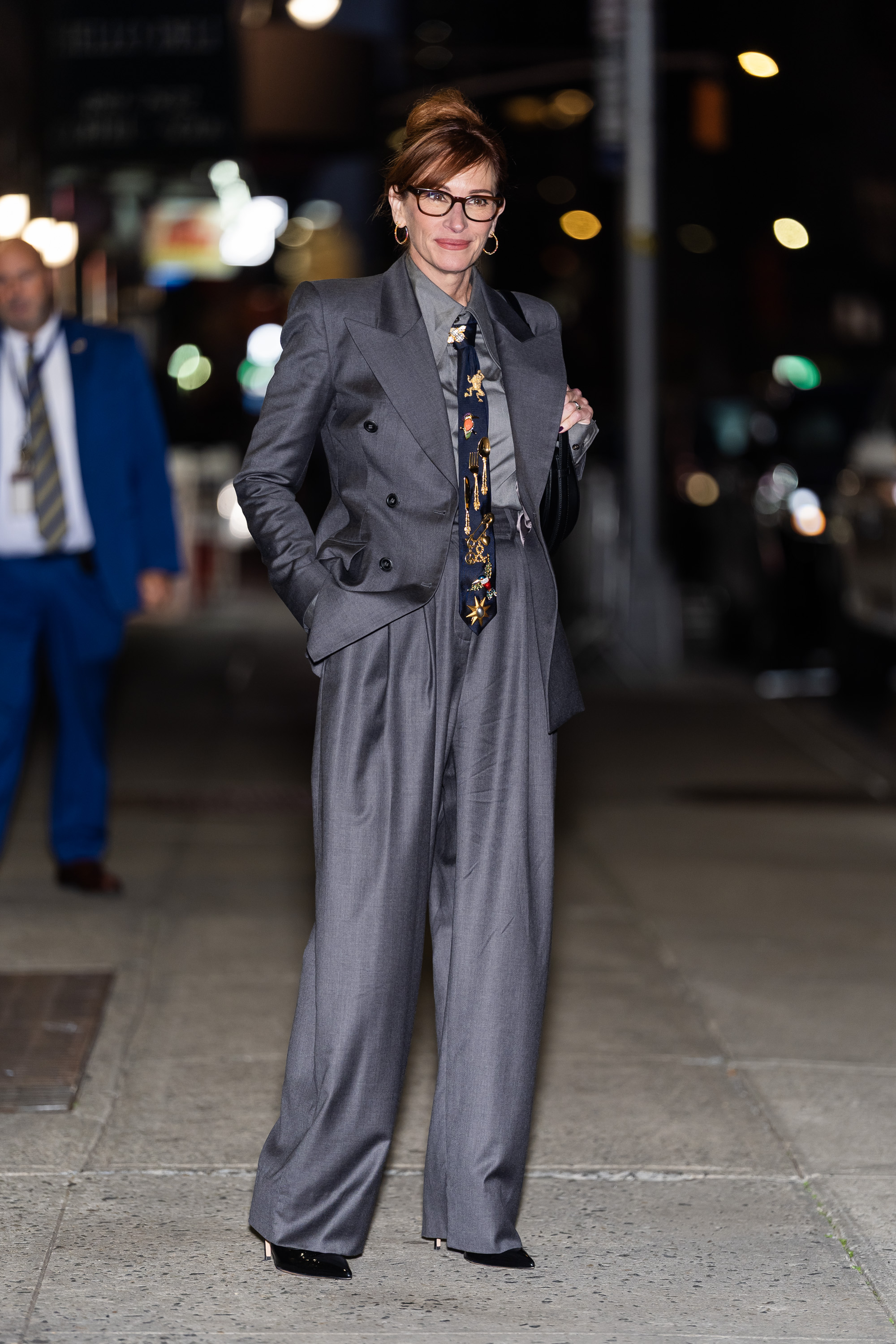 An image of actor Julia Roberts wearing a charcoal grey suit with a light grey shirt, a navy tie with various pins, heels and black-framed glasses