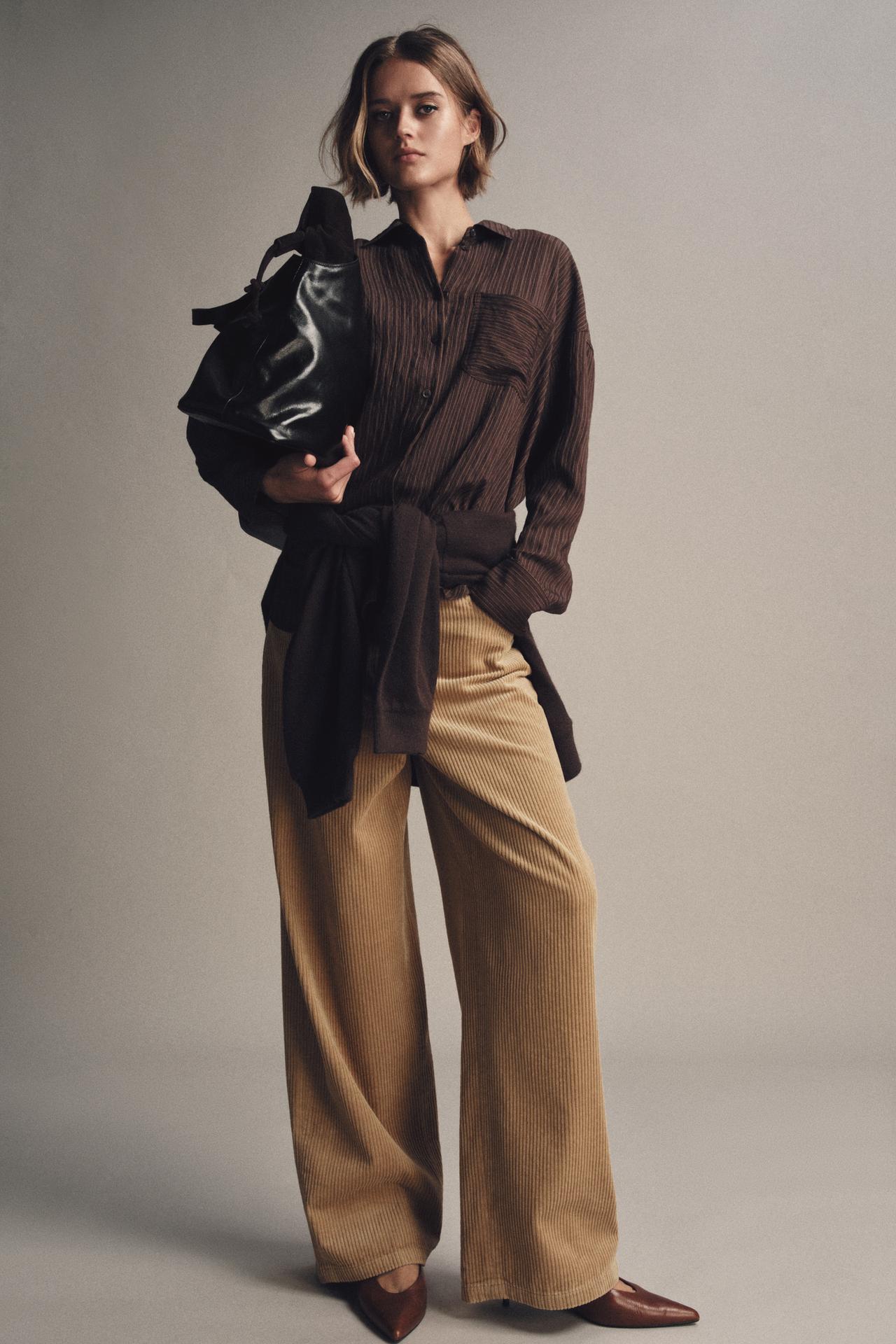 model wears brown striped button dow, tan corduroy pants, brown pointed toe kitten heels, and black tote with a sweatshirt around her waist
