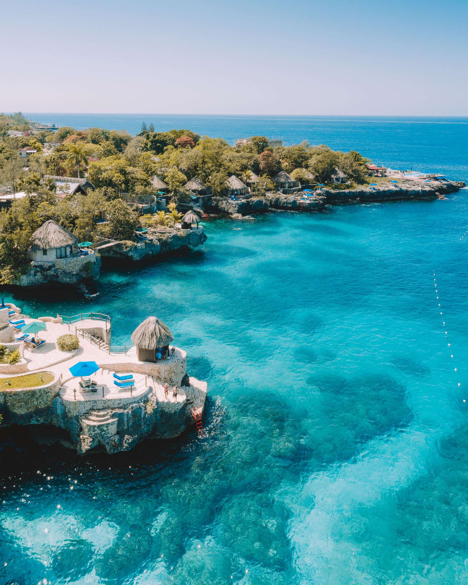 The Rockhouse Hotel in Jamaica pictured from an areal view. Pictured is a bright blue clear sea with little huts dotted along the cliff side with red ladders that dip into the sea. A few blue umbrellas and lounge chairs are along the edge as well.