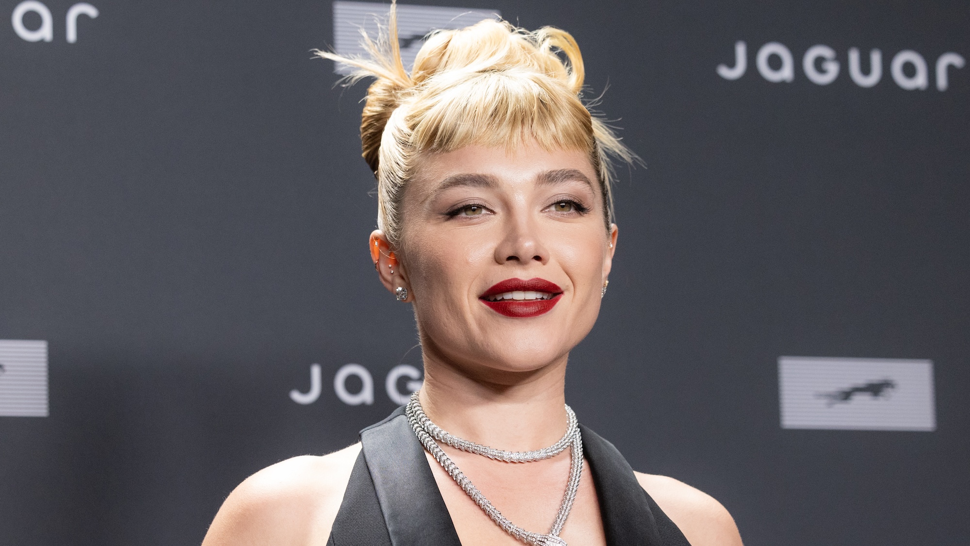 Florence Pugh smiling with red lipstick and updo at the Jaguar event in 2026.