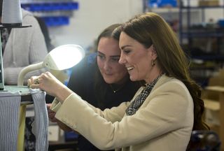 Kate Middleton wearing a white coat and plaid tie-neck dress sitting in front of a machine holding a pair of blue socks as a woman watches on