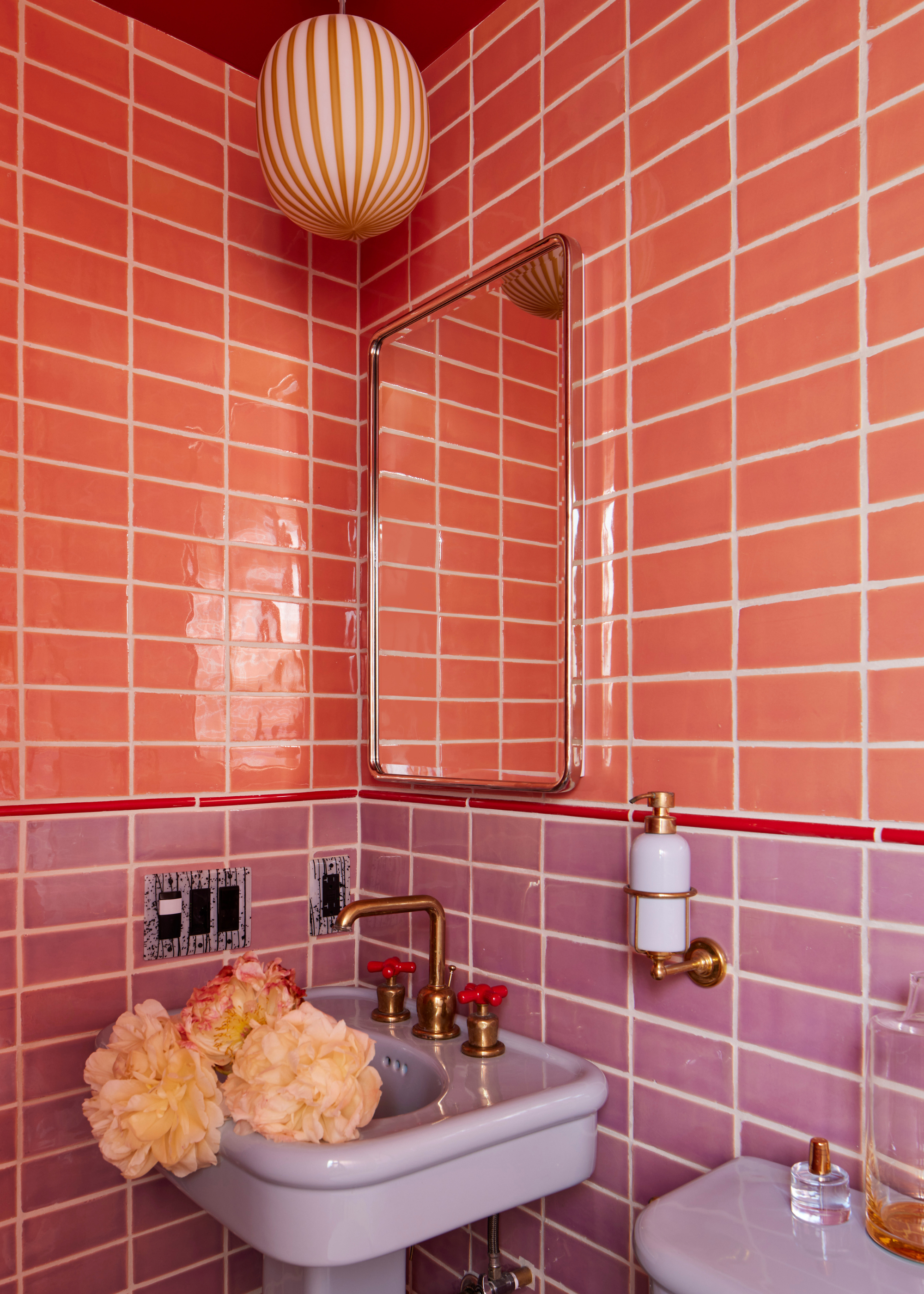 A small bathroom with dual tile drenching in a coral pink and soft purple. There is a curved mirror on the wall, cut flowers in the sink and brass hardware on the sink and the wall-attached soap dispenser