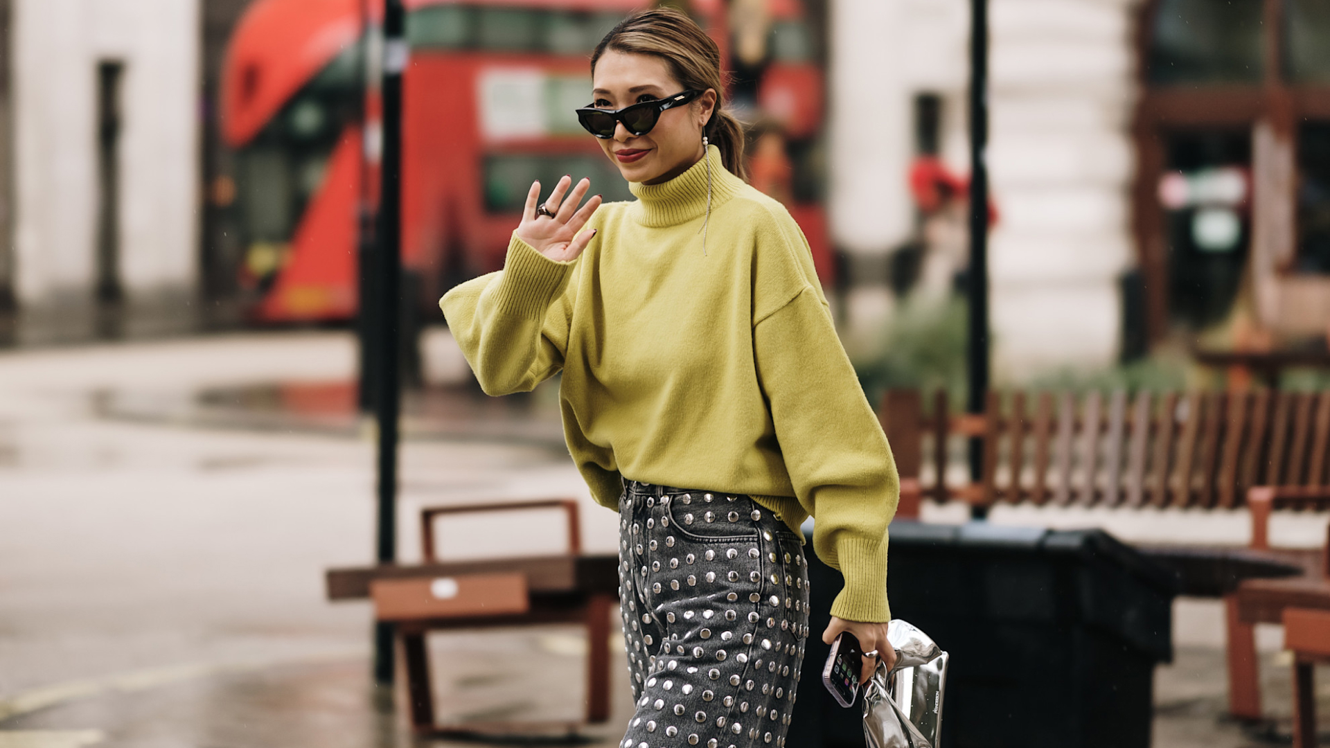 fashion week attendee waving wearing sunglassess, green sweater, and studded jeans