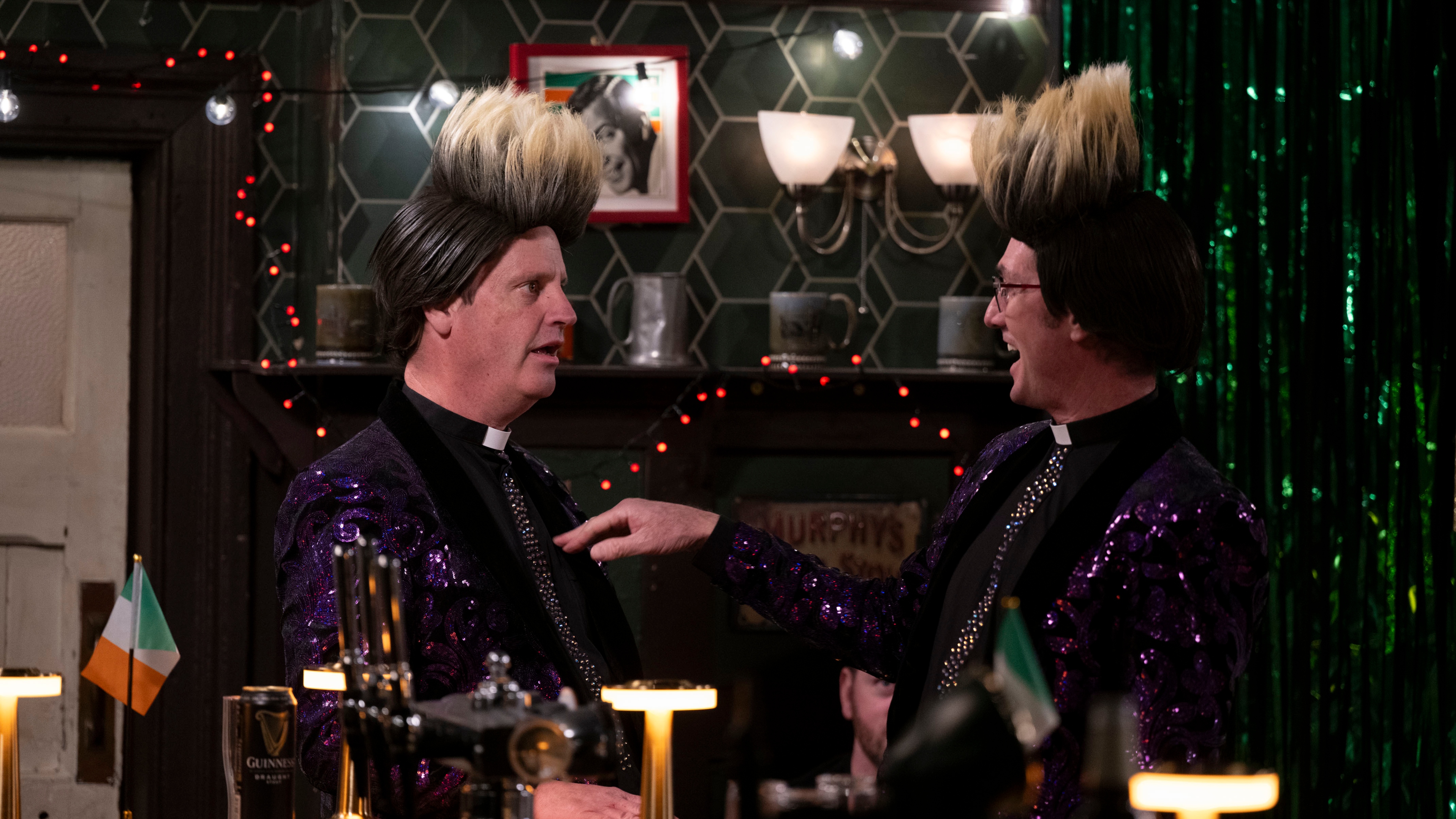 Fathers Damien (Conor Moloney) and Trevor (Martin Delany) performing as Jedward with large blond wigs and sequenced purple jackets in Foley's Bar.