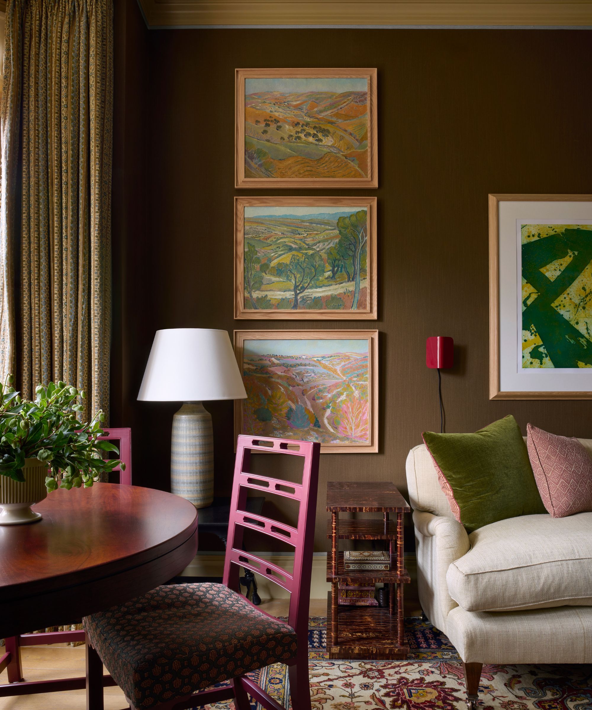 Brown sitting room with antique paintings on the wall, round antique table with pink dining chairs, cream sofa, green and pink pillows, a Persian rug, and a bamboo side table.