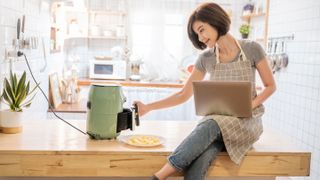 A woman cooking with an air fryer with a laptop balanced on her lap while sitting on a counter