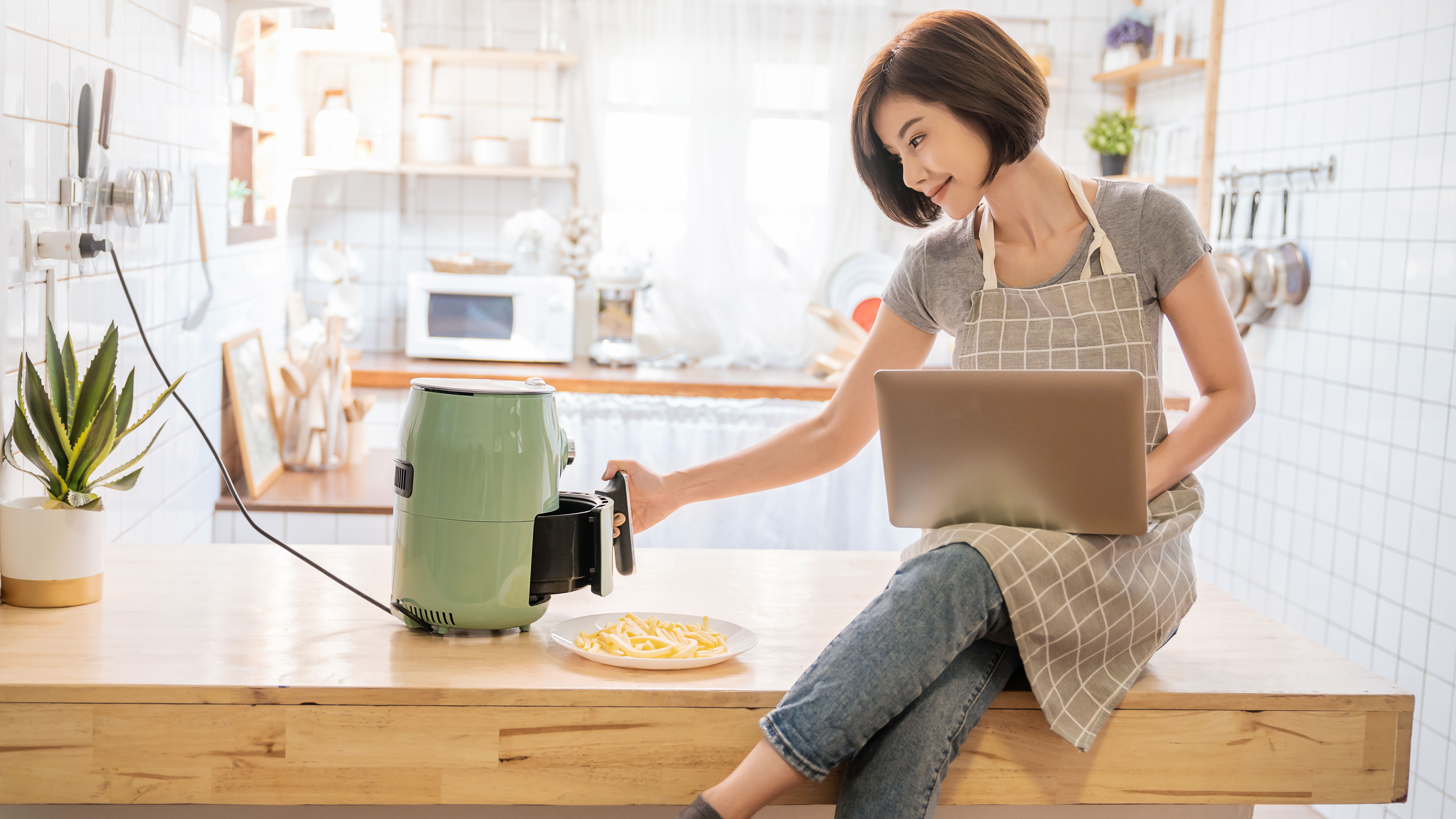 Woman cooking with an air fryer with laptop balanced on her lap while sitting at a table
