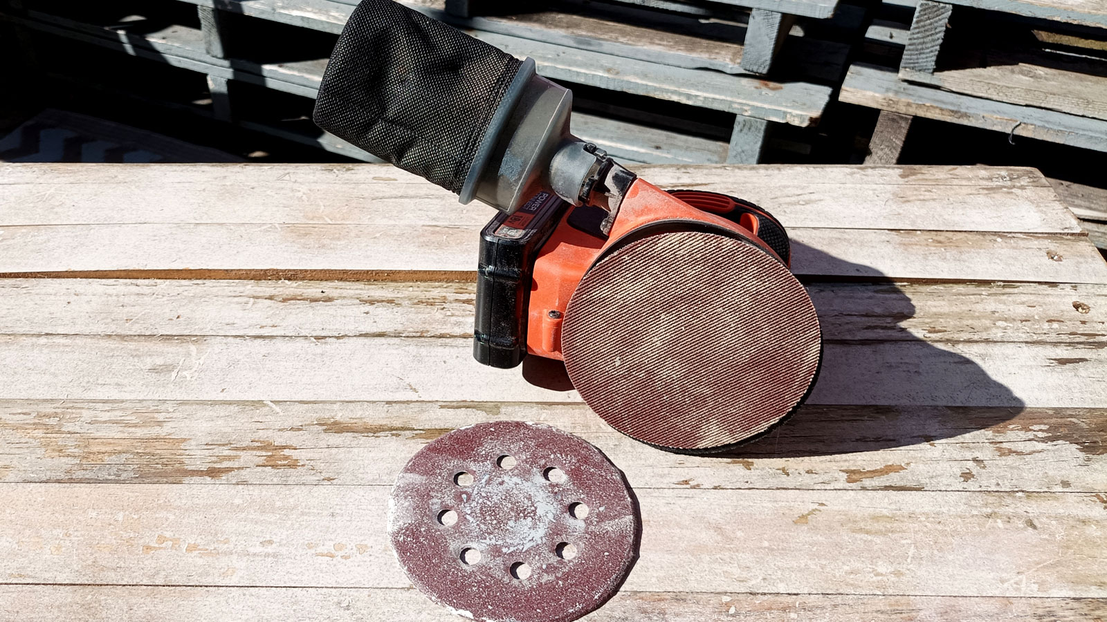 Orange orbital sander on its side with a mesh sanding disc on the sander and a paper-based disc with holes in front