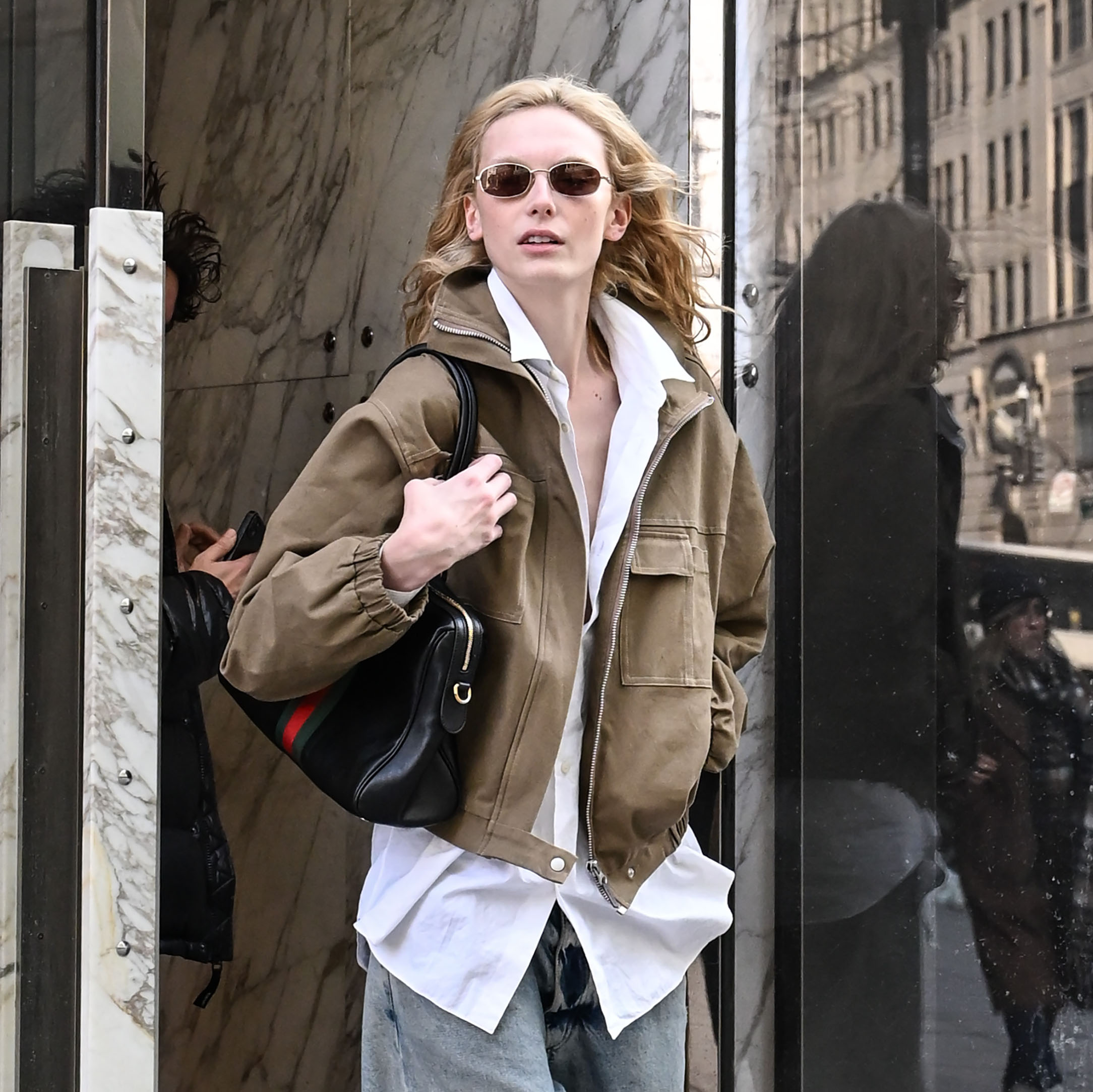 Alex Consani wearing a brown jacket, white button-down, blue jeans, black loafers, and black shoulder bag