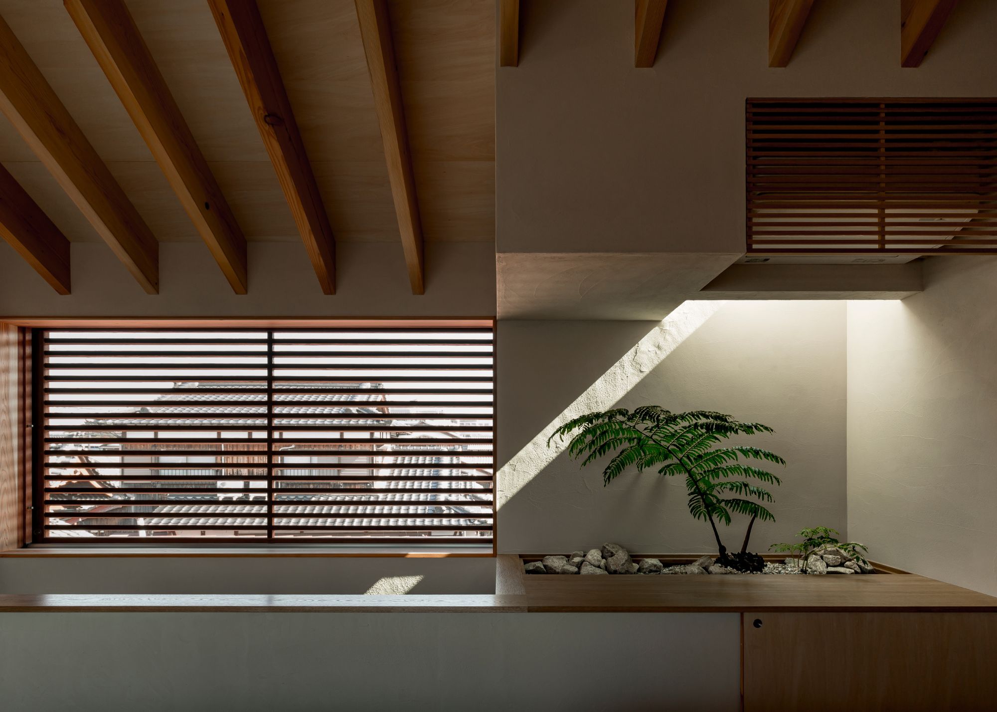 A minimalist Japanese space with wood beams, shutters and details, and a solitary plant in the corner in amongst stones and pebbles comes to life by the beam of natural light just above it