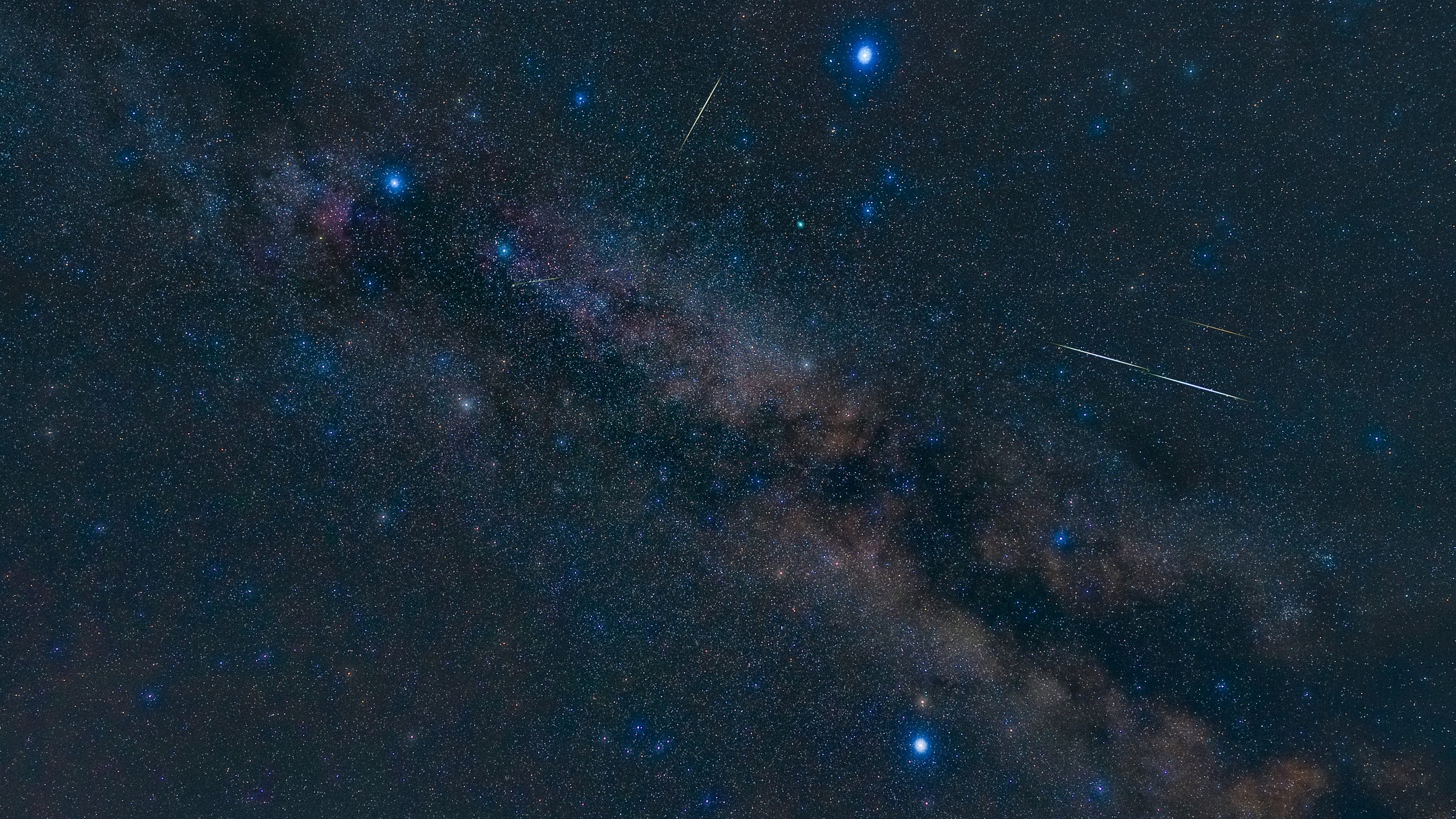 drie heldere sterren zijn gerangschikt in een driehoekige formatie aan de nachtelijke hemel.  Sommige meteoorstrepen zijn zichtbaar over de afbeelding en de Melkweg strekt zich uit over de achtergrond.