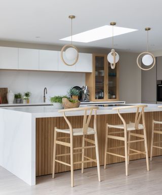 A white waterfall kitchen island with a wooden base, three Scandi chairs in front of it, three round pendant lights above it, and white cabinets behind it