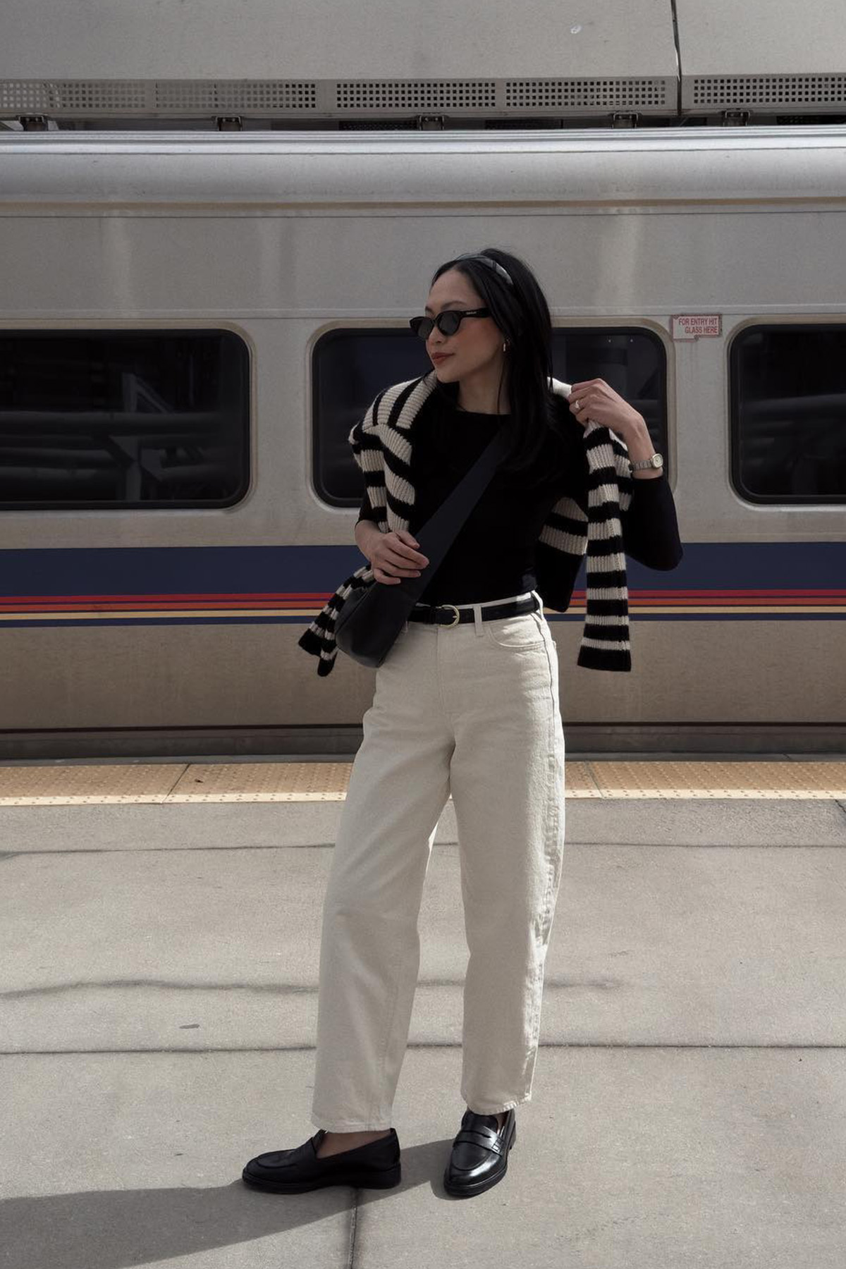 How to style white jeans for fall, shown in a photo of a woman standing outside at a train station wearing a black headband, black sunglasses, a black boatneck top with a striped sweater over the shoulders, a black belt, and white baggy jeans, and black loafers