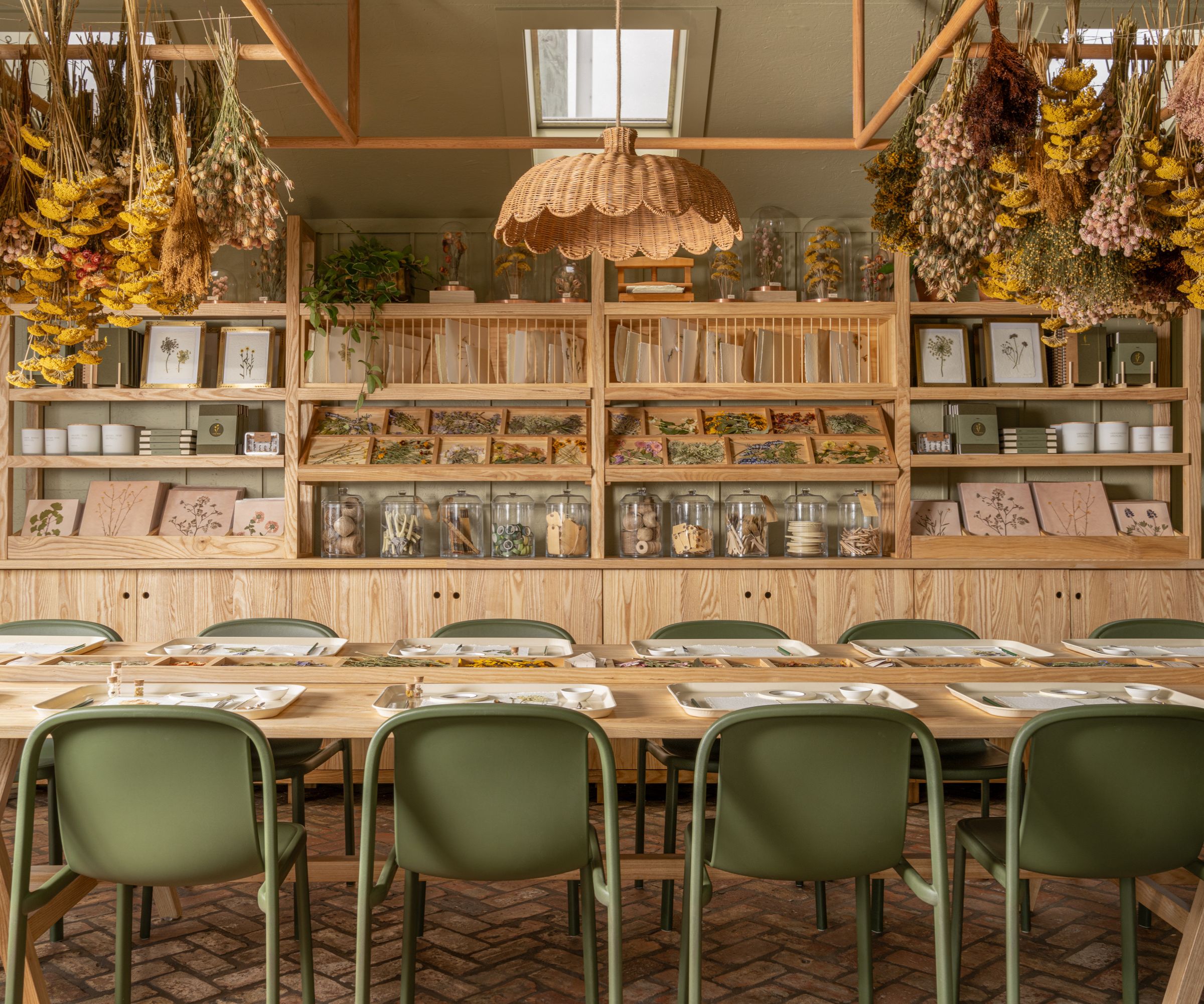 Room with brick floor, sage green painted walls, dried flowers hanging on ceiling, rattan light and long wood dining table with green chairs