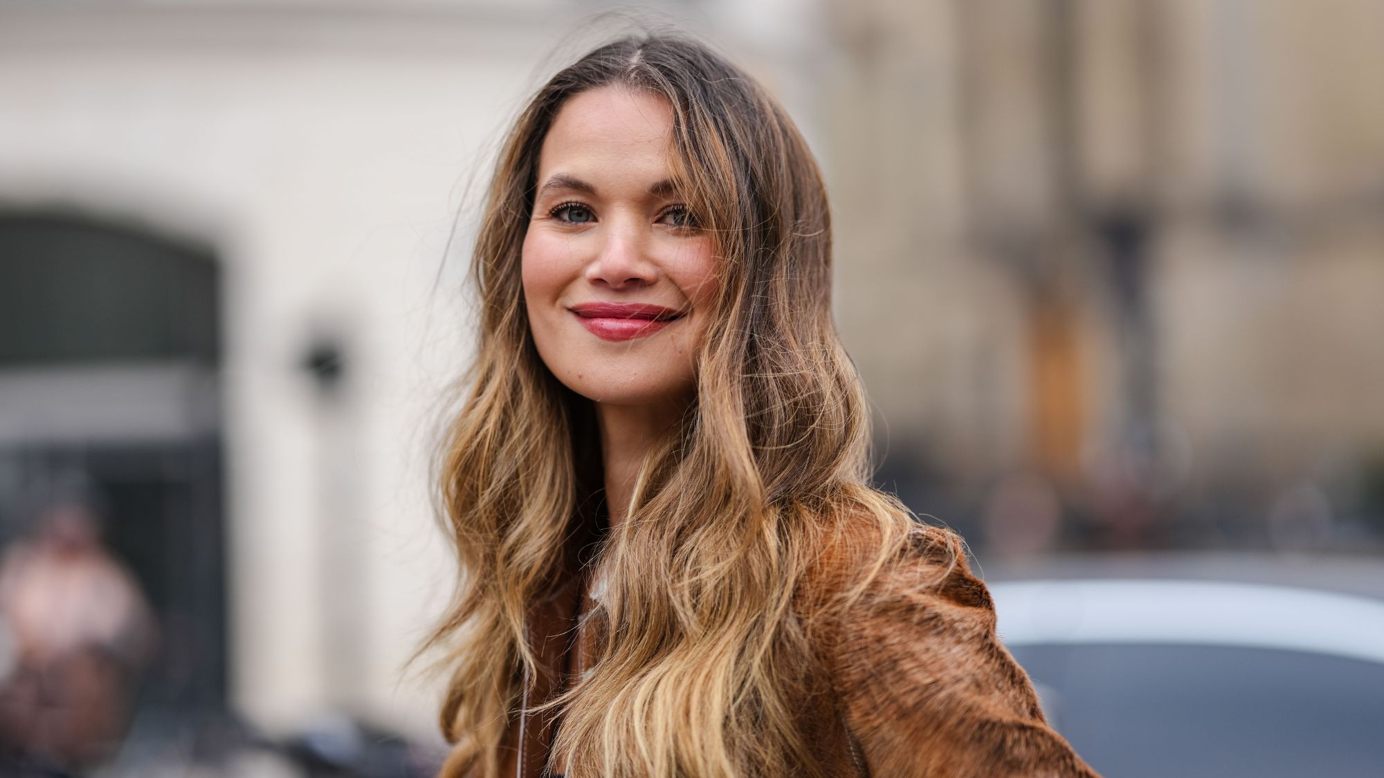 street style shot of woman with long wavy hair and pink lipstick smiling