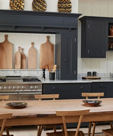 A kitchen with dark painted cabinets, marble countertops, wooden table, chairs and cutting boards, a silver cooker and white tiles with yellow and black decorative plates above