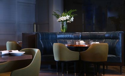 Interior view of Disgruntled Chef featuring grey panelled walls and a door, a dark coloured curved seat, dining tables with tableware, muted green chairs and a clear vase with white flowers and foliage