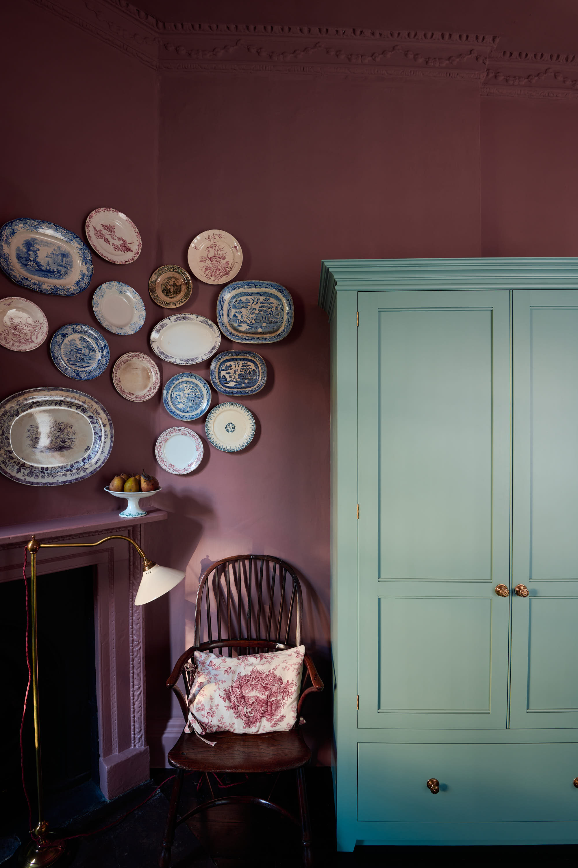 a room with deep red painted walls, a mint green cabinet, a chair with a pillow, and a cluster of plates hanging in the corner of the wall