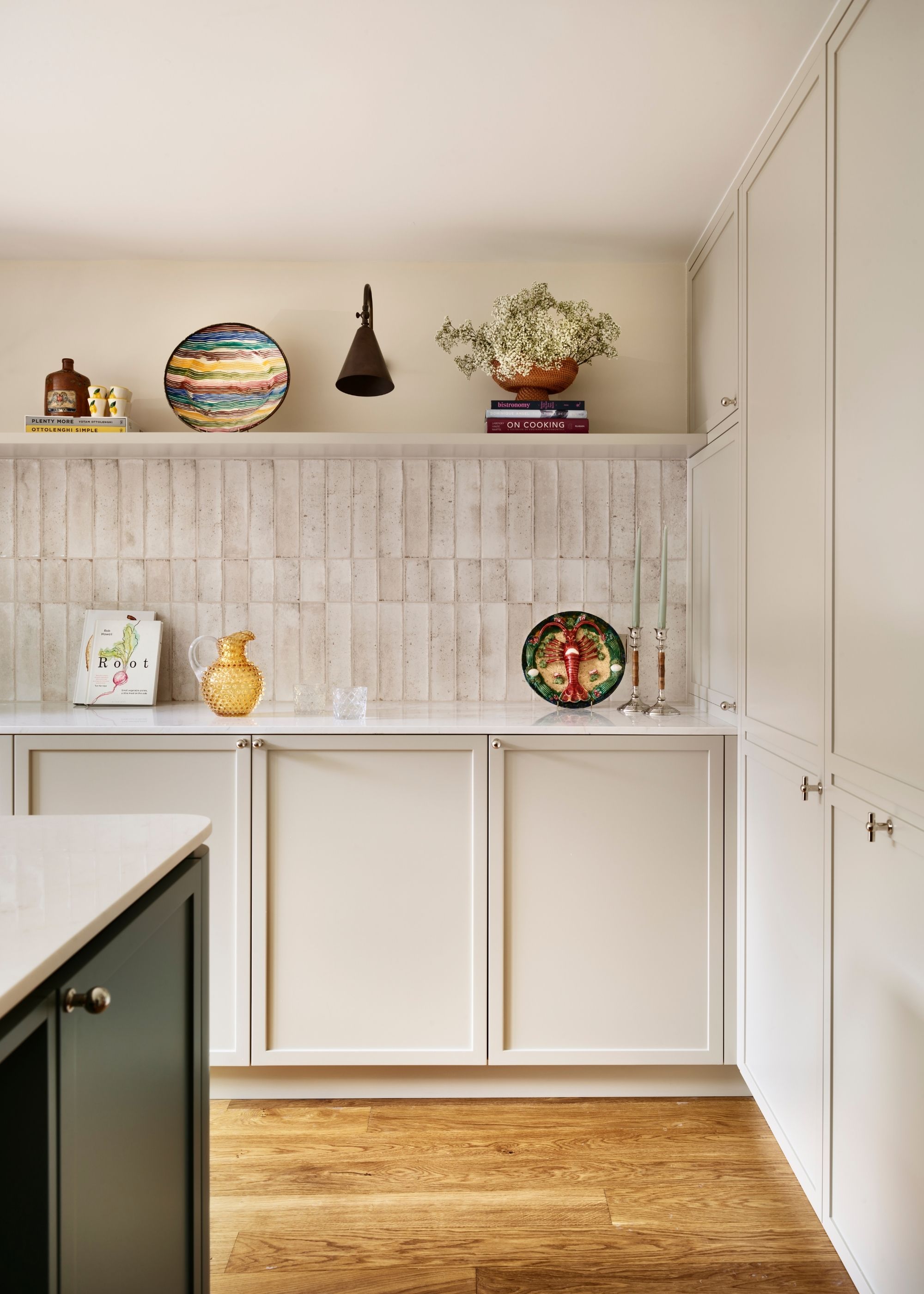 A modern kitchen with white neutral cabinets, white gloss wall tiles, oak flooring, and decorative objects on a shelf.
