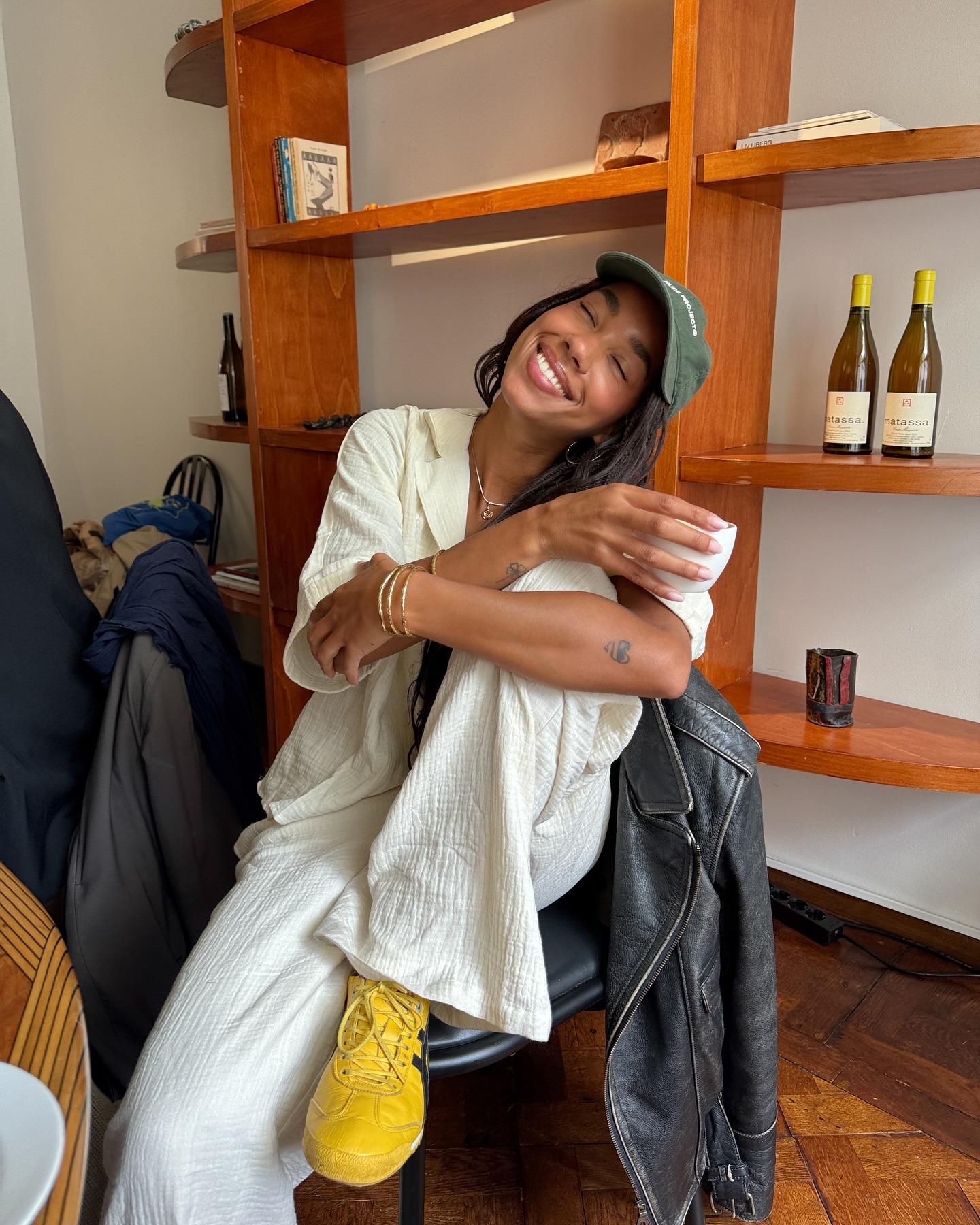 Fashion person Amaka Hamelijnck wears a linen set, green cap and Onitsuka tiger yellow trainers. She is holding a cup of coffee, sat a on a chair in a coffee shop smiling into the camera in a recent image from her Instagram.