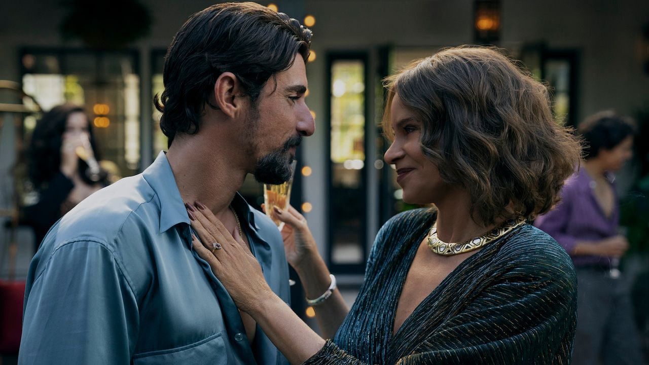 a man (Alberto Guerra as Dario) gazes at a woman (Sofia Vergara as Griselda) as she places a hand on his chest and holds a glass of champagne during a party