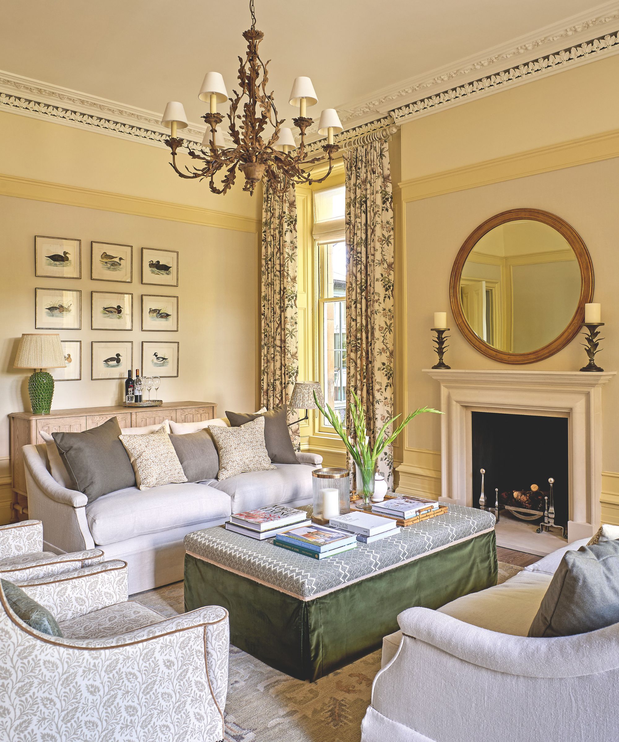 Pale yellow traditional living room with circular mirror, gray sofas, green ottoman and stone fireplace
