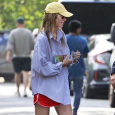 Olivia wilde wears red athletic shorts, a yellow baseball cap, and a button-down shirt.