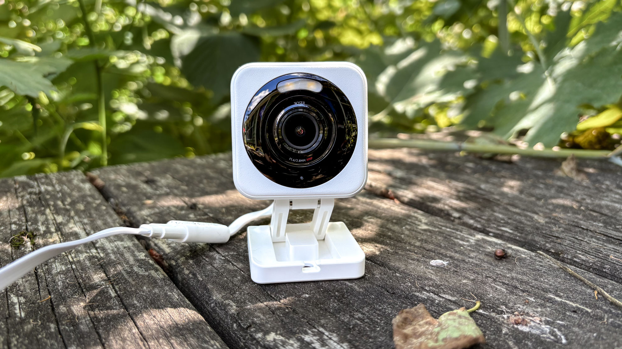 A Wyze security camera outside on a deck in front of green bushes.