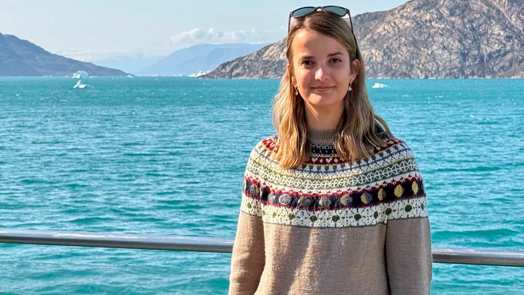 a woman in a sweater smiles for a portrait on the deck of a cruise ship in arctic waters, with a mountain range visible in the distance