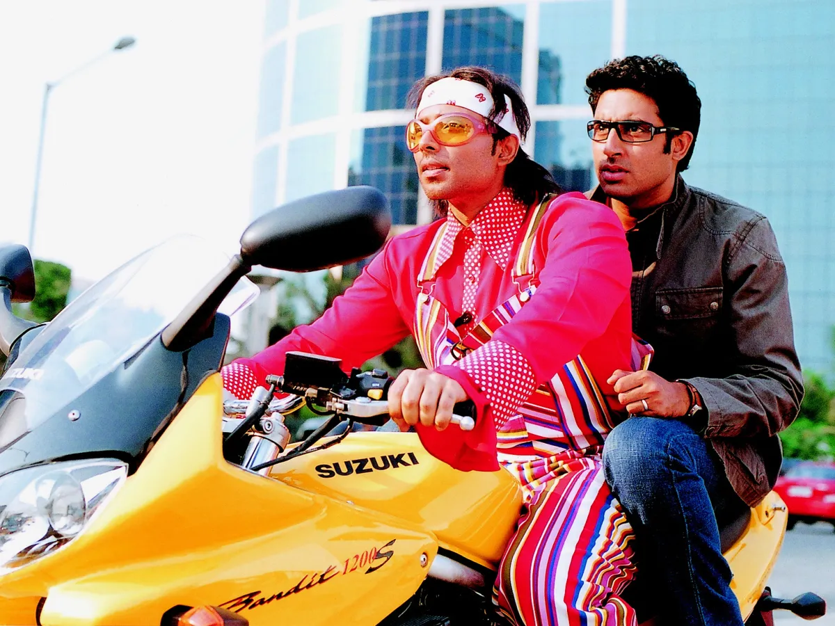 a man in a pink shirt and striped overalls and a man in jeans ride on. yellow bike together in a still from dhoom